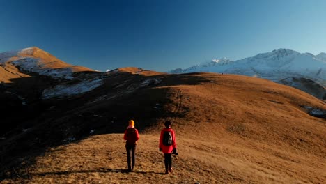 Aerial-view-of-a-two-girls-traveler-with-backpacks-and-cameras-stroll-through-the-hills-between-the-epic-rocks-in-the-mountains.-Girls-photographers-with-their-cameras-at-sunset