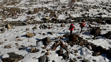 Luftaufnahme-von-zwei-Mädchen-Reisenden-mit-Rucksäcken-und-Kameras-gehen-durch-den-Schnee-und-Steinen-auf-den-Gletscher-zwischen-den-epischen-Felsen-in-den-Bergen.-Mädchen-Fotografen-mit-ihren-Kameras-bei-Sonnenuntergang