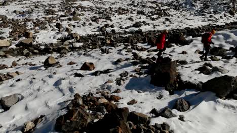 Vista-aérea-del-viajero-dos-chicas-con-mochilas-y-cámaras-ir-por-la-nieve-y-las-piedras-para-el-glaciar-entre-las-rocas-épicas-en-las-montañas.-Fotógrafos-de-las-niñas-con-sus-cámaras-al-atardecer