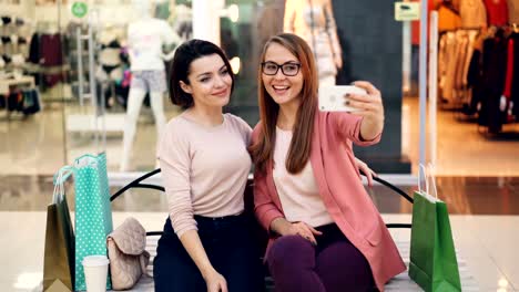 Gut-aussehende-junge-Damen-nehmen-Selfie-zusammen-mit-Smartphone-sitzen-im-Einkaufszentrum-ausruhen-und-Spaß-haben.-Mädchen-sind-umarmt-und-posiert-mit-Handgesten.