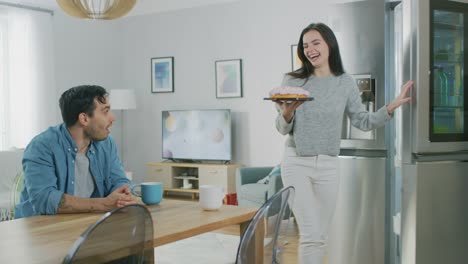 Hermosa-joven-pareja-hablando-en-la-cocina-en-casa.-Hombre-está-sentado-en-una-mesa,-la-chica-toma-un-pastel-de-la-nevera.-THAY-están-felices-y-ríen.-Habitación-tiene-un-Interior-moderno.