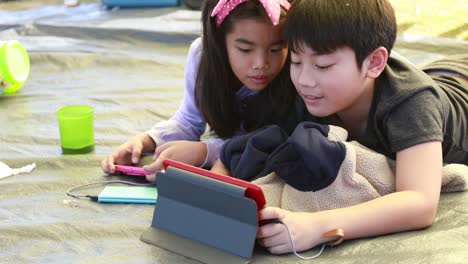 Happy-asian-boy-and-girl-playing-smart-phone-and-tablet-in-camping-area