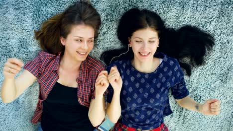 Top-view-of-two-beautiful-girls-in-headphones-listening-music-dancing-and-smiling-while-lying-on-bed