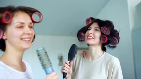 Dos-chicas-divertidas-cantando-con-peines-bailan-y-tienen-alegría-en-la-sala-de-estar-en-casa