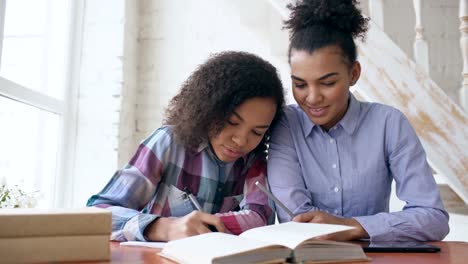 Teenage-curly-haired-mixed-race-young-girl-sitting-at-the-table-concentrating-focused-learning-lessons-and-her-elder-sister-helps-her-studying