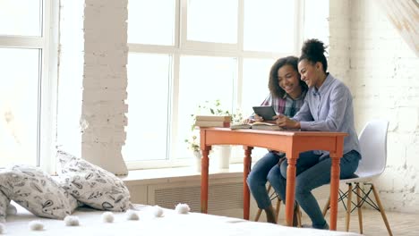 Two-attractive-curly-haired-mixed-race-young-girls-sitting-at-the-table-have-fun-while-learning-lessons-and-using-tablet-at-home