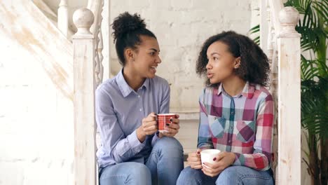 Two-african-american-curly-girls-sistres-sitting-on-stairs-have-fun-laughing-and-chatting-together-at-home