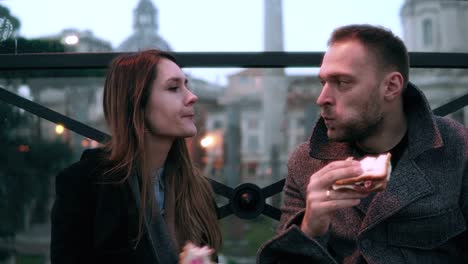 Young-stylish-couple-sitting-at-the-city-centre-and-eating-sandwiches-in-evening.-Looks-aside-together