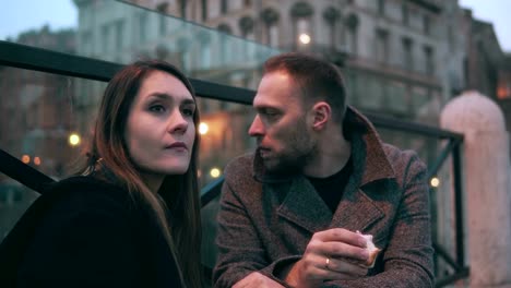 Young-man-and-woman,-two-friends-sitting-on-the-bridge-in-evening-and-eating-sandwiches.-Couple-have-a-break