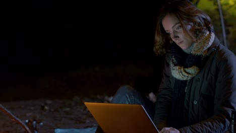 Brunette-girl-is-using-a-laptop-next-to-a-campfire-at-night-with-a-tent-in-the-background.