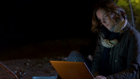 Brunette-Mädchen-benutzt-nachts-einen-Laptop-neben-einem-Lagerfeuer-mit-einem-Zelt-im-Hintergrund.