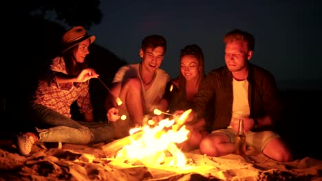 Junge-fröhliche-Freunde-abends-am-Feuer-am-Strand-sitzen,-Kochen-Eibisch-auf-zusammen-klebt.-Schuss-in-4k