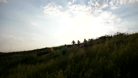 Gruppe-junger-Männer,-die-den-grünen-Hügel-hinunter-über-blauer-Himmel-mit-Sonne-Streulicht-im-Hintergrund-laufen.-Männliche-Athleten-ist-Joggen-in-der-Natur-bei-Sonnenuntergang.-Kufen-bergab-Sport-im-Freien-bei-Sonnenaufgang.-Slow-motion