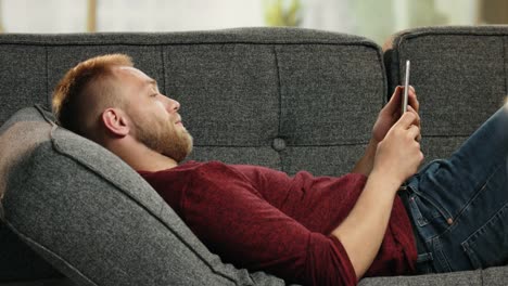 Atractivo-joven-acostado-en-el-sofá-leyendo-noticias-divertidas-sobre-tableta