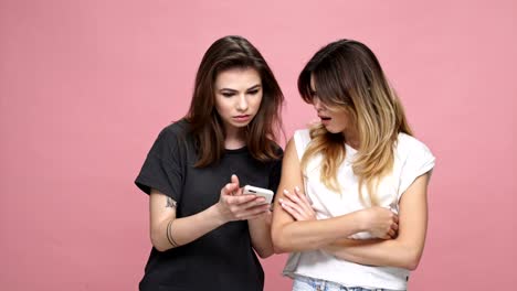 Two-young-casual-girls-looking-at-mobile-phone-and-getting-upset-isolated-over-pink-background