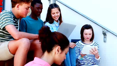 Group-of-smiling-school-friends-on-staircase-using-laptop-and-digital-tablet