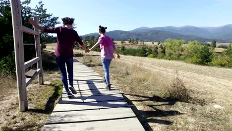 Dos-expresivas-chicas-divirtiéndose-en-el-paseo-del-parque-de-montaña-jugar-avión-vuelo.-Están-saltando-emocionados,-vista-posterior