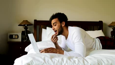 Pondering-Man-Using-Laptop-Computer-Lying-On-Bed-Hispanic-Guy-Type-Chatting-Online-In-Bedroom-Morning