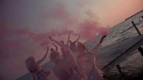 Multi-ethnic-friends-celebrating-with-smoke-bombs-on-jetty-at-sunset