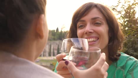 Two-happy-young-woman-tourists-sitting-at-the-table-of-a-bar-restaurant-in-front-of-the-Colosseum-in-Rome-drink-and-toast-with-a-glass-of-italian-red-wine.-Stylish-colorful-dress-on-a-summer-day-at-sunset