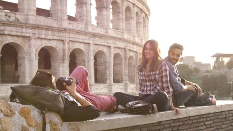 Mochilas-de-tres-turistas-jóvenes-amigos-sentados-mentira-delante-de-Coliseo-de-Roma-al-atardecer-tomando-fotos-fotos-con-dslr-cámara-gafas-de-sol-feliz-hermosa-chica-de-cabello-largo-lenta