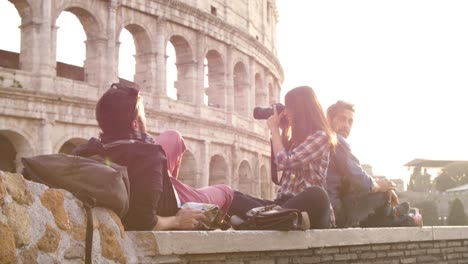 Mochilas-de-tres-turistas-jóvenes-amigos-sentados-mentira-delante-de-Coliseo-de-Roma-al-atardecer-tomando-fotos-fotos-con-dslr-cámara-gafas-de-sol-feliz-hermosa-chica-de-cabello-largo-lenta