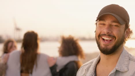 Atractivo-hombre-sonriente-a-la-cámara-mientras-que-las-mujeres-busca-buenas-bailando-en-el-fondo,-al-atardecer