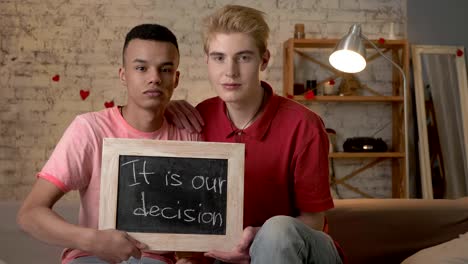 A-sad-international-gay-couple-is-sitting-on-the-couch-and-holding-a-sign.-It-is-our-decision.-Look-at-the-camera.-Home-comfort-on-the-background.-60-fps