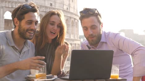 Drei-glückliche-Freunde-mit-Videoanruf-mit-Laptop-bar-Restauranttisch-vor-Colosseum-in-Rom-bei-Sonnenuntergang-winkenden-Händen-geben-Daumen-nach-oben-zu-sitzen