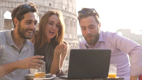 Three-happy-friends-having-video-call-with-laptop-sitting-at-bar-restaurant-table-in-front-of-colosseum-in-rome-at-sunset-waving-hands-giving-thumbs-up