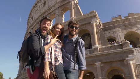 Three-young-friends-tourists-standing-on-pedestal-in-front-of-colosseum-in-rome-taking-funny-hilarious-pictures-posing-with-backpacks-sunglasses-happy-beautiful-girl-long-hair-slow-motion