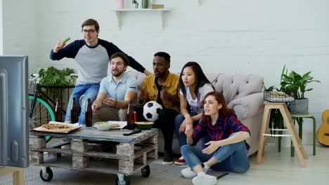 Group-of-young-friends-watching-football-game-on-TV-together-eating-snacks-and-drinking-beer-at-home.-Men-are-happy-with-their-team-winning-but-girls-disappointed