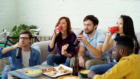 Amigos-felices-han-partido-hablando-y-comiendo-pizza.-Ellos-clink-sentado-en-el-sofá-en-casa-de-botellas