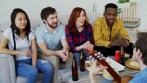 Happy-young-friends-have-party-at-shared-apartment-and-singing-together-while-their-friend-playing-guitar-at-home