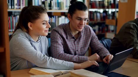 Alegre-y-universitarios-trabajando-en-ordenador-portátil-junto-sentado-en-la-mesa-en-la-biblioteca-de-la-Universidad-con-compañeros-de-clase-sobre-fondo