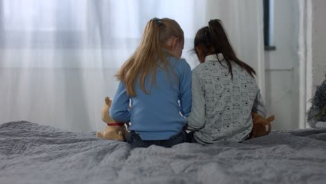 Back-view-little-multi-ethnic-girls-sitting-on-bed