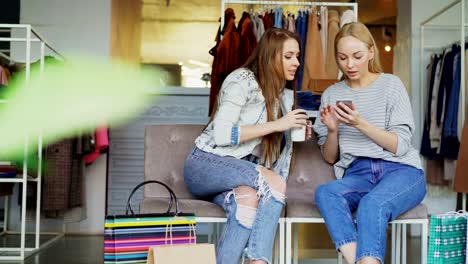 Amigas-hablando-y-control-smartphone-sentado-es-centro-comercial.-Ropa-y-bolsas-de-papel-en-fondo-de-las-mujeres.