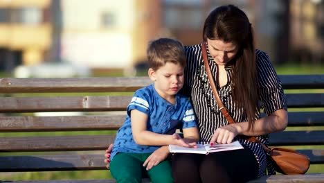 Joven-madre-y-su-hijo-sentados-en-un-banco-de-un-parque-y-leer-un-libro