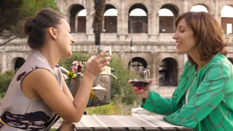 Two-happy-young-woman-tourists-sitting-at-the-table-of-a-bar-restaurant-in-front-of-the-Colosseum-in-Rome-drink-and-toast-with-a-glass-of-italian-red-wine.-Stylish-colorful-dress-on-a-summer-day-at-sunset