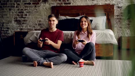 Cute-young-couple-boyfriend-and-girlfriend-are-playing-videogame-holding-joysticks-sitting-on-bedroom-floor-at-home.-Emotional-lovers-are-enjoying-game.