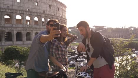 Drei-glückliche-junge-Freunde-Touristen-mit-Fahrrädern-und-Rucksäcke-am-Kolosseum-in-Rom-unter-Selfies-auf-Hügel-bei-Sonnenuntergang-mit-Bäumen-Zeitlupe-steadycam