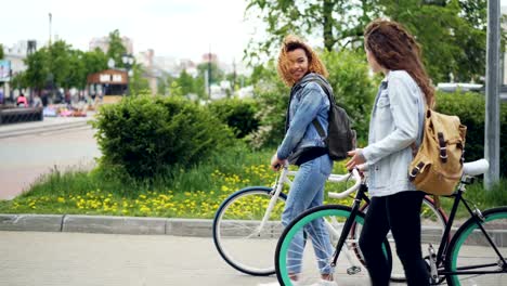 Afroamerikanische-Frau-spaziert-mit-ihrem-kaukasischen-Freund-halten-Fahrrad-entlang-der-schönen-Straße-mit-Brunnen-und-Holzbänken,-Bäume-und-Autos-sind-sichtbar.