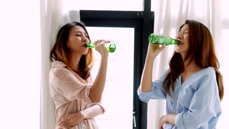 Young-asian-young-woman-lesbian-couple-standing-clinking-bottles-of-beer-at-home.