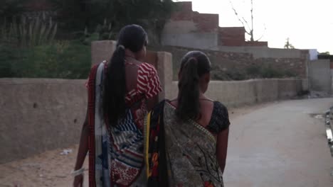 Two-girls-walk-stroll-holding-hands-friends-bond-home-streets-rural-setting-happy-bonding-share-cute--outdoor-meet-follow-gimbal-behind-village-small-town-India-traditional-dress-costume-brick-wall