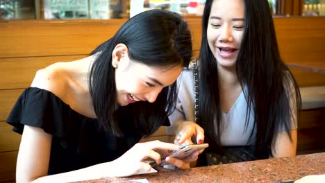 Dos-asiáticas-amigas-usando-teléfono-móvil-y-riendo-juntos.-Mujeres-disfrutando-de-buen-tiempo-en-el-restaurante-japonés