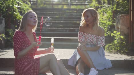 Two-attractive-beautiful-young-blond-women-blow-bubbles-in-the-park-and-are-happy-on-sunset.-Girlfriends-in-park-on-the-steps