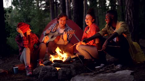 Cinemagraph-loop---cheerful-guy-in-casual-clothing-is-playing-the-guitar-while-his-male-and-female-friends-tourists-are-singing-and-smiling-sitting-around-fire-in-forest.