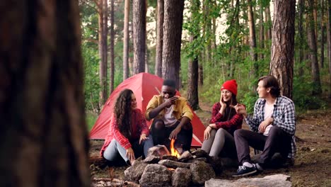 Cheerful-girls-and-guys-hikers-are-talking-and-laughing-sitting-near-fire-at-campsite-sharing-stories-and-having-fun.-Green-trees,-modern-tent-and-flame-are-visible.