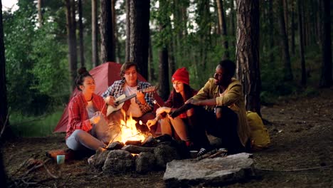 Turístico-es-tocar-la-guitarra-sentado-junto-a-la-fogata-con-amigos-cantando-y-divirtiéndose,-jóvenes-tienen-palillos-con-malvavisco-por-encima-de-la-llama.-Concepto-de-comida,-música-y-diversión.