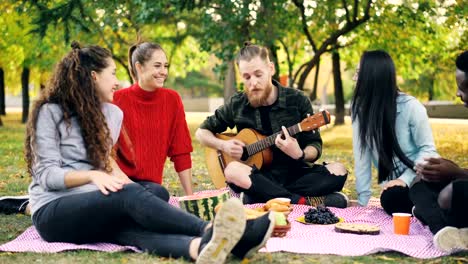 Chico-guapo-es-tocar-la-guitarra-mientras-su-hombre-y-amigas-escuchando-música,-cantando-y-divirtiéndose-en-el-picnic-en-el-parque.-Concepto-de-amistad-y-cultura.